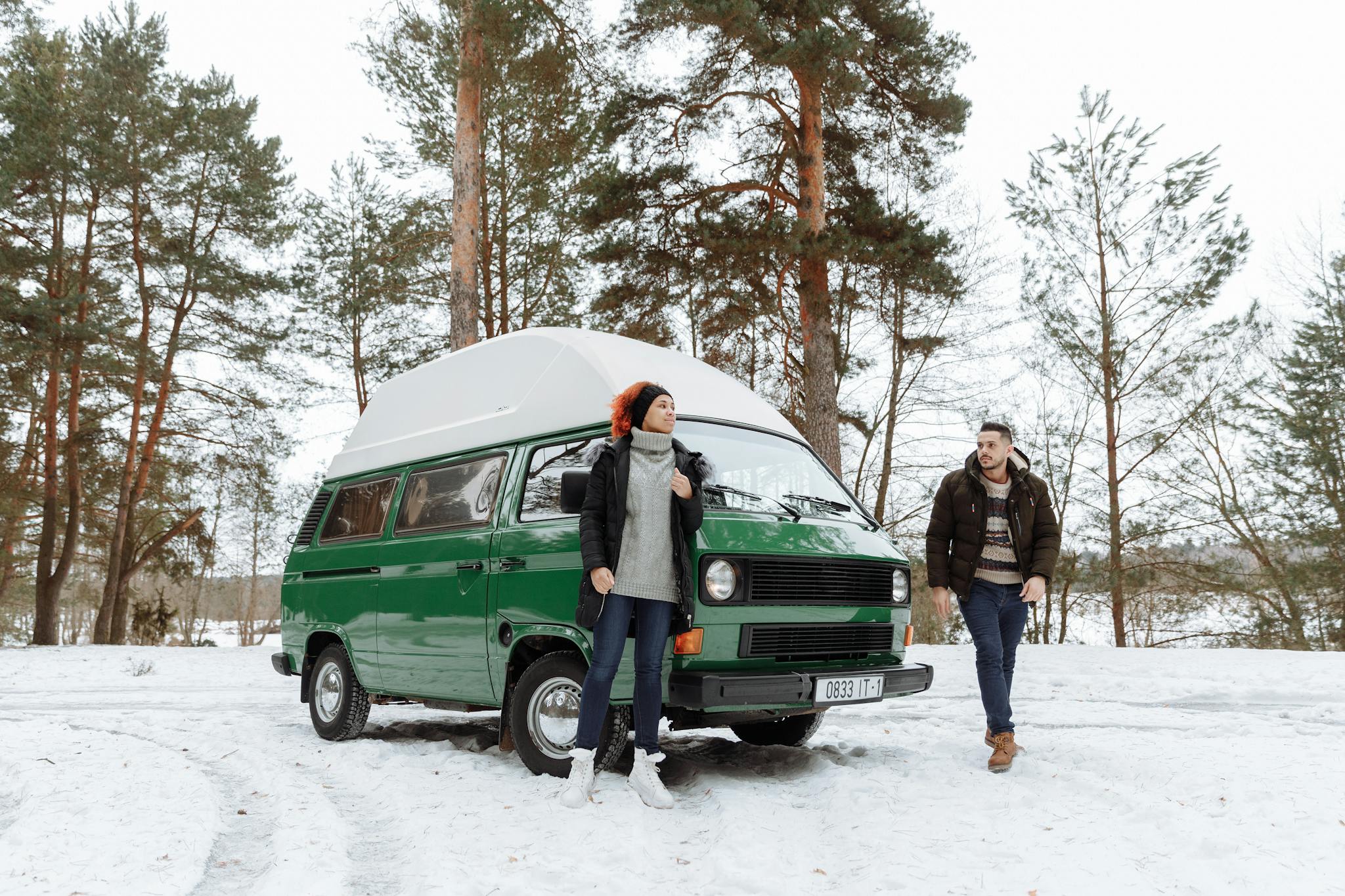 A couple enjoys a winter adventure with their green campervan in a snowy forest.